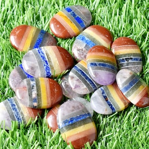 May include: A collection of seven smooth, oval-shaped stones, each layered with seven different colored bands of semi-precious stones. The stones are arranged on a bed of green grass.
