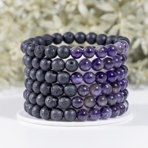 May include: A stack of beaded bracelets. The bracelets alternate between black lava stone and purple amethyst beads. The beads are round and smooth. The bracelets are stacked on a white surface, with a blurred background of white and green.