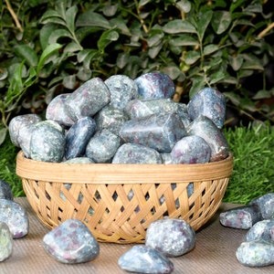 Puede incluir: Una cesta de mimbre llena de piedras pulidas de color azul grisáceo con inclusiones rojas. Algunas piedras están esparcidas alrededor de la cesta sobre una superficie neutra, con follaje verde en el fondo. Las piedras tienen una forma lisa y redondeada.