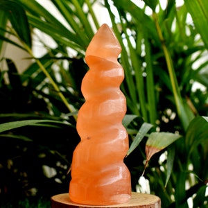 May include: A spiral-shaped pink Himalayan salt crystal, carved into a tower shape. The crystal is sitting on a wooden base.