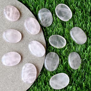 May include: Oval rose quartz stones in varying shades of pink. The polished stones are displayed on a green and tan surface. The stones are smooth and reflect light. The image shows a collection of these stones.