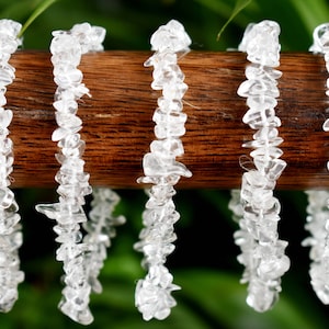 Puede incluir: Cinco pulseras de chips de cristal de cuarzo transparente apiladas sobre una superficie de madera. Las pulseras están hechas de pequeños trozos irregulares de cuarzo que están enhebrados en un elástico.