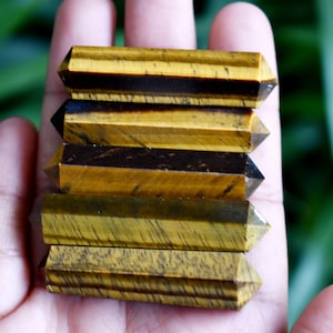 May include: A stack of five golden tiger's eye crystal points, each with a unique pattern of brown and gold stripes. The points are arranged in a pyramid shape, with the largest point at the bottom and the smallest point at the top.