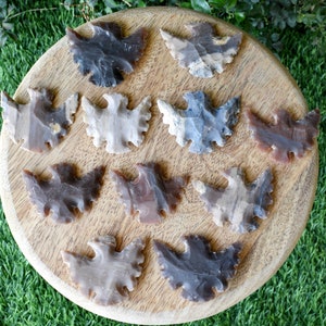 May include: A collection of 12 brown, grey, and white arrowhead-shaped stones with a bird design carved into them. The stones are arranged on a wooden surface.