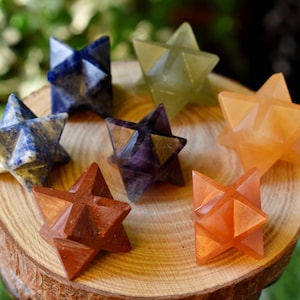 Könnte beinhalten: Sieben sternförmige Kristallspitzen, jede in einer anderen Farbe, angeordnet in einem Kreis auf einer Holzoberfläche. Die Farben sind blau, grün, orange, rot, lila und zwei Orangetöne.