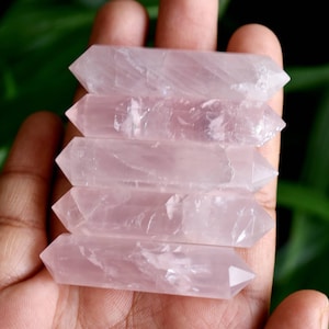 May include: Five polished rose quartz crystal wands, each with pointed ends, are held in a hand. The crystals have a soft pink hue and a translucent appearance. The background is a blurred green, suggesting foliage.