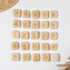 May include: Twenty-four beige rune stones, each with a gold symbol, arranged on a white surface. The stones are rectangular with rounded corners. The runes are in a grid pattern. A wooden decorative element is in the background.