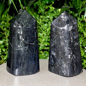 Puede incluir: Dos obeliscos de turmalina negra con acabado pulido. Los cristales tienen un aspecto oscuro, casi opaco, con sutiles vetas y motas de color más claro. Se exhiben sobre una superficie neutra contra un fondo de follaje verde.