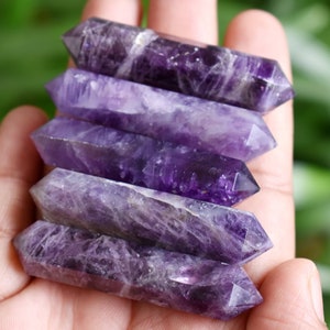 May include: Five amethyst crystal wands, each with a double-pointed tip, are stacked on top of each other in a hand. The wands are a deep purple color with white streaks throughout.