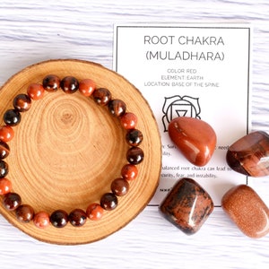 May include: Red and black beaded bracelet on a wooden coaster with four red and brown stones. The stones are associated with the root chakra, also known as Muladhara. The card reads "ROOT CHAKRA (MULADHARA) COLOR RED ELEMENT EARTH LOCATION BASE OF THE SPINE".