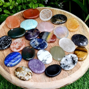 May include: A collection of 20 smooth, oval-shaped polished stones, each with a unique color and pattern, arranged in a circle on a wooden surface. The stones include amethyst, rose quartz, clear quartz, tiger's eye, and more.