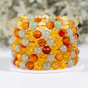 May include: Stack of beaded bracelets in shades of orange, yellow, and green. The bracelets are made of round beads, with variations in color and transparency. The bracelets are stacked on a white surface, creating a visually appealing display.