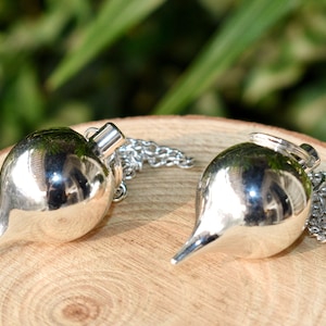 Op de afbeelding: Twee zilverkleurige pendels met een druppelvorm en een kleine ketting. De pendels liggen op een houten oppervlak met een vage groene achtergrond. De pendels hebben een glanzend, reflecterend oppervlak.