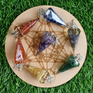 Könnte beinhalten: Sieben Kristall-Pendel mit silbernen Ketten, die in einem Kreis auf einem Holzsockel mit geometrischem Muster angeordnet sind. Die Pendel bestehen aus klarem Harz, das mit verschiedenen Kristallen gefüllt ist, darunter Amethyst, Karneol und grüner Aventurin.
