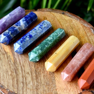 Könnte beinhalten: Sieben polierte Edelstein-Zauberstäbe in verschiedenen Farben, darunter Lila, Blau, Grün, Gelb, Rosa und Rot, liegen auf einer Holzoberfläche. Die etwa 10 cm langen Stäbe haben facettierte Enden, die die Farben hervorheben.