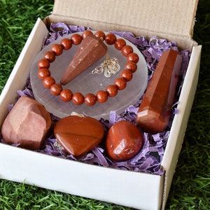 May include: A white gift box filled with red jasper crystals. The box contains a red jasper bracelet, a red jasper heart pendant, a red jasper point, a red jasper tumbled stone, and a red jasper tower. The box is sitting on a bed of green grass.