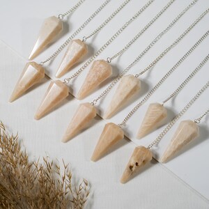 Op de afbeelding: Een verzameling perzikkleurige kristallen pendulums met zilveren kettingen. Elke pendel heeft een facetvormige, kegelachtige vorm. De pendulums zijn diagonaal geplaatst op een witte ondergrond, met wat dorre grassprieten in de linker benedenhoek.