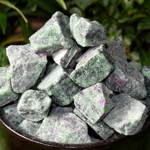 May include: A pile of green and black rough zultanite gemstones. The stones are in a black ceramic bowl.