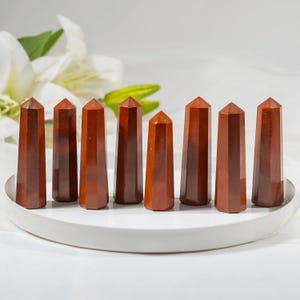 以下が含まれることがあります： さまざまな高さの研磨された赤い碧玉のオベリスクが7つ、白い丸いトレイに配置されています。オベリスクは多面的なデザインで、豊かな赤茶色をしています。背景には白い花と緑の葉が含まれています。