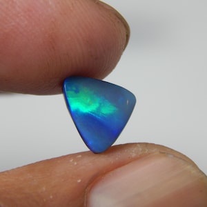 Puede incluir: Una gema de ópalo triangular pulida. La piedra muestra un vibrante juego de colores, con destellos verdes y azules. La gema se sostiene entre dos dedos sobre un fondo blanco liso.