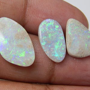 Peut inclure: Trois pierres d'opale avec un éclat nacré. Les pierres présentent un mélange de couleurs blanches, vertes et bleues. Les opales sont de formes variées, dont ovales et en forme de goutte, sur fond neutre.