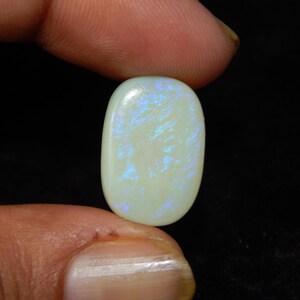 May include: An oval-shaped opal gemstone, polished to a smooth finish. The opal displays a creamy white base with iridescent flashes of blue and green. The gemstone is held between fingers against a black backdrop.