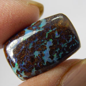 May include: A polished, rectangular gemstone with a dark brown base and intricate patterns of blue, green, and iridescent colors. The stone is held between two fingers, showcasing its unique, natural beauty.
