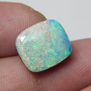 May include: A square-cut opal gemstone with a mix of colors, including blue, green, and white. The opal has a textured surface and a polished appearance. The gemstone is held between fingers.