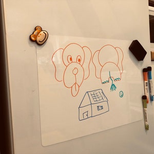 Puede incluir: Una pizarra blanca magn&eacute;tica de borrado en seco con dibujos de una casa, un perro y un elefante. Hay un im&aacute;n de mono en la esquina superior izquierda de la pizarra. Tambi&eacute;n hay tres marcadores de colores en el lado derecho de la pizarra.