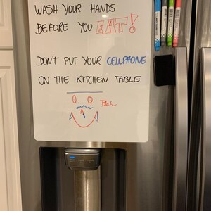 Puede incluir: Una pizarra blanca de borrado en seco con el texto "WASH YOUR HANDS BEFORE YOU EAT!" y "DON'T PUT YOUR CELLPHONE ON THE KITCHEN TABLE" escrito con un marcador negro. Hay cinco marcadores de borrado en seco de colores diferentes unidos a la pizarra con una tira magn&eacute;tica negra.