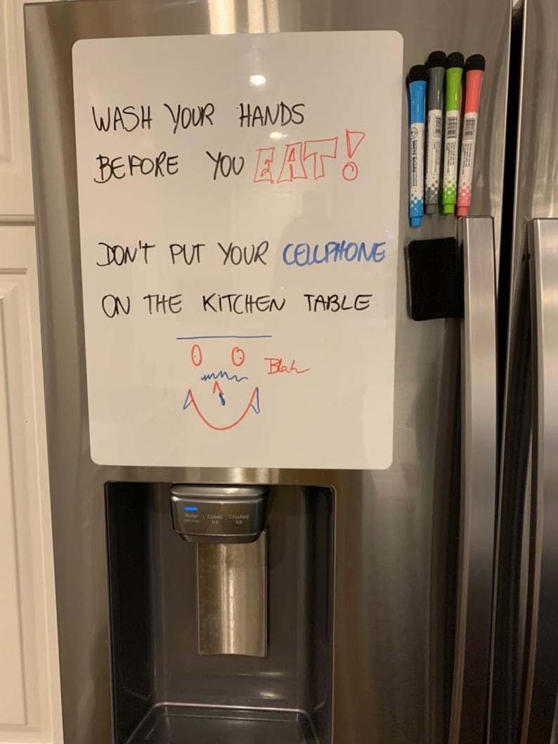 Puede incluir: Una pizarra blanca de borrado en seco con el texto "WASH YOUR HANDS BEFORE YOU EAT!" y "DON'T PUT YOUR CELLPHONE ON THE KITCHEN TABLE" escrito con un marcador negro. Hay cinco marcadores de borrado en seco de colores diferentes unidos a la pizarra con una tira magn&eacute;tica negra.