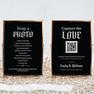 May include: Two black framed signs with gold trim. The left sign says "Snap a PHOTO" and lists photo ideas for a wedding. The right sign says "Capture the LOVE" and has a QR code to share photos. The signs are on a white surface with a gold mesh overlay.