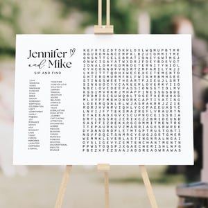 May include: A white rectangular sign featuring the text Jennifer and Mike Sip and Find, a list of wedding-themed words, and a large word search puzzle grid containing various romantic terms printed in black ink on a white background.