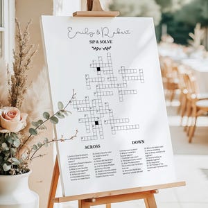 May include: A white wedding crossword puzzle sign on a wooden easel featuring the names Emily & Robert, the title SIP & SOLVE, and numbered clues for across and down. The sign is displayed next to a white vase containing dried pampas grass and a rose.