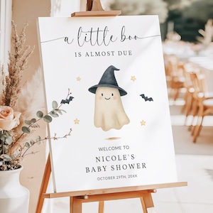 以下が含まれることがあります： 木製のイーゼルに立てられた白い看板。テキストは「a little boo is almost due」と「Welcome to Nicole's Baby Shower」。魔女の帽子をかぶった漫画の幽霊、コウモリ、星が描かれています。日付は20XX年10月29日です。