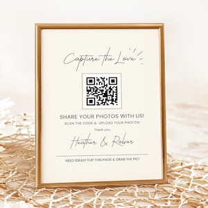 May include: A gold-framed sign with a white background and black text. The sign says "Capture the Love" and has a QR code. The text below the QR code says "Share your photos with us! Scan the code & upload your photos! Thank you. Heather & Rober. Need ideas? Flip this page & grab the pic!"