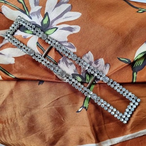 May include: A silver-toned rectangular buckle with a double row of clear rhinestones. The buckle is lying on a brown fabric with a floral pattern.