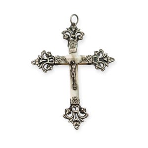 Op de afbeelding: Een zilverkleurige metalen crucifix hanger met een centrale inleg van wit parelmoer. Het kruis heeft sierlijke, decoratieve metalen filigraan uiteinden en een kleine metalen figuur van Jezus in het midden, met de letters INRI erboven gegraveerd.