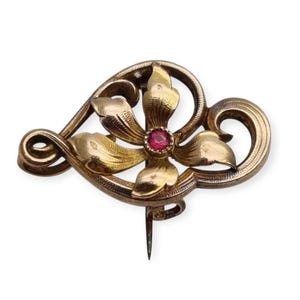 Puede incluir: Un broche dorado en forma de corazón con un diseño de flor estilizado. La flor tiene una piedra preciosa roja en el centro.
