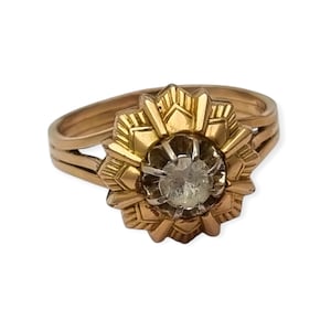 Puede incluir: Un anillo de oro con diseño floral. El anillo presenta una piedra preciosa central y transparente rodeada por un engaste de oro en forma de flor. La banda se divide en dos hebras paralelas. El anillo está sobre un fondo blanco.