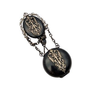 Può includere: Una collana con pendente antico argento e nero. La collana presenta due pendenti neri ovali e circolari con ornati disegni in filigrana argentata. I pendenti sono collegati da una catena d'argento.