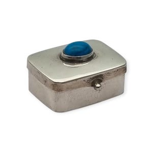 May include: A rectangular silver-toned metal box with a smooth, polished lid featuring a single, centered, oval-shaped turquoise blue cabochon stone. The box has a small, rounded knob on the front edge for opening the hinged lid.