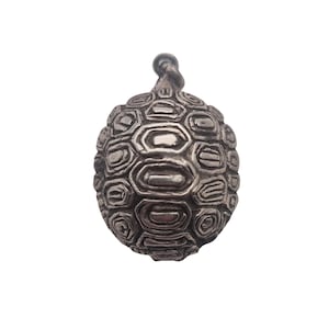 May include: A silver-toned metal pendant shaped like a turtle shell, featuring a detailed, raised geometric pattern of hexagonal and pentagonal plates across its rounded surface, with a small circular loop at the top for attachment to a chain.