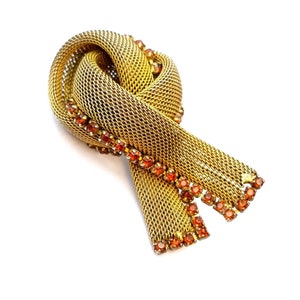 May include: A gold mesh brooch with a knotted design and orange rhinestones.