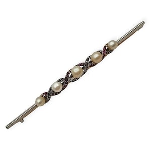 May include: A long, slender bar brooch featuring five round, cream-colored pearls set in a diagonal, twisted metal frame. The frame is accented with small, deep red gemstones and tiny clear stones, creating a textured, interwoven pattern along the center.