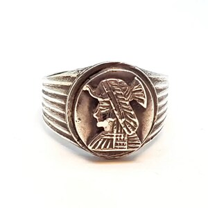 May include: A silver ring with a raised design of a woman's profile wearing a helmet. The ring has a textured band.