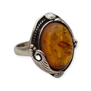 Puede incluir: Anillo ovalado de ámbar con montura de metal plateado. El anillo presenta una piedra de ámbar con un tono marrón dorado cálido, engastada en un marco decorativo con detalles de hojas y rayos de sol. El anillo es una joya.