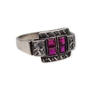 Puede incluir: Un anillo de tono plateado con cuatro piedras de color rosa vibrante de corte cuadrado dispuestas en un grupo central. La banda está decorada con filas de pequeñas piedras redondas oscuras en los bordes y piedras claras en los lados.