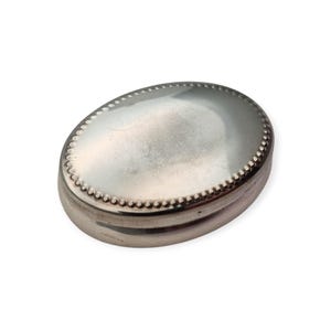 May include: An oval-shaped silver-toned metal box with a smooth, reflective lid surface. The perimeter of the lid features a decorative beaded border detail. The base of the container is solid and matches the metallic finish of the lid.