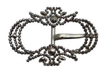 May include: A silver-toned metal buckle with a decorative design of small beads and stones. The buckle is oval-shaped and has a simple metal bar closure.